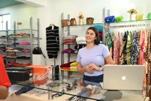 A diferença entre MEI e ME na prática é ilustrada por uma jovem empreendedora sorridente atrás do balcão de vidro de sua loja de roupas, acompanhada de um notebook e caixas de produtos.