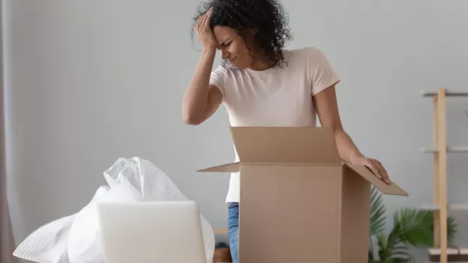 Uma mulher está ao lado de uma mesa, segurando a cabeça em frustração enquanto olha para uma caixa de papelão aberta. Ao lado dela está um laptop e alguns materiais de embalagem, sugerindo que ela está desempacotando ou embalando itens. O fundo possui uma estante parcialmente montada.